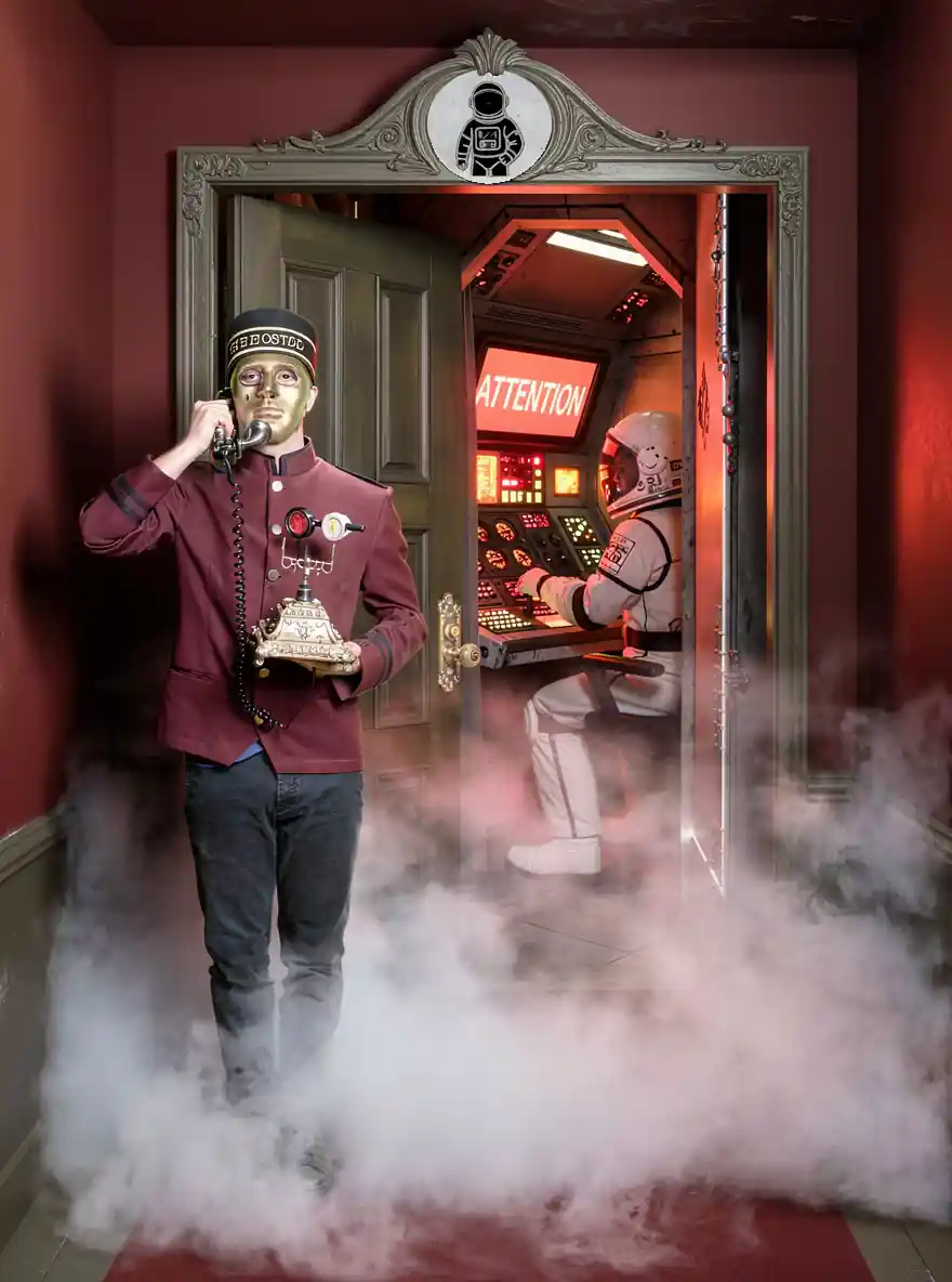 Un majordome en uniforme rouge et masque doré, parle dans un téléphone ancien devant une porte ouverte révélant un astronaute assis à un tableau de contrôle rétro éclairé en rouge avec le mot 'ATTENTION'. Une légère brume entoure leurs pieds, ajoutant une ambiance mystérieuse