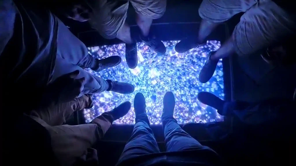 Image de plusieurs paires de pieds et posées sur une surface lumineuse, bleutée et scintillante, comme un sol interactif ou une vitrine éclairée dans une ambiance sombre