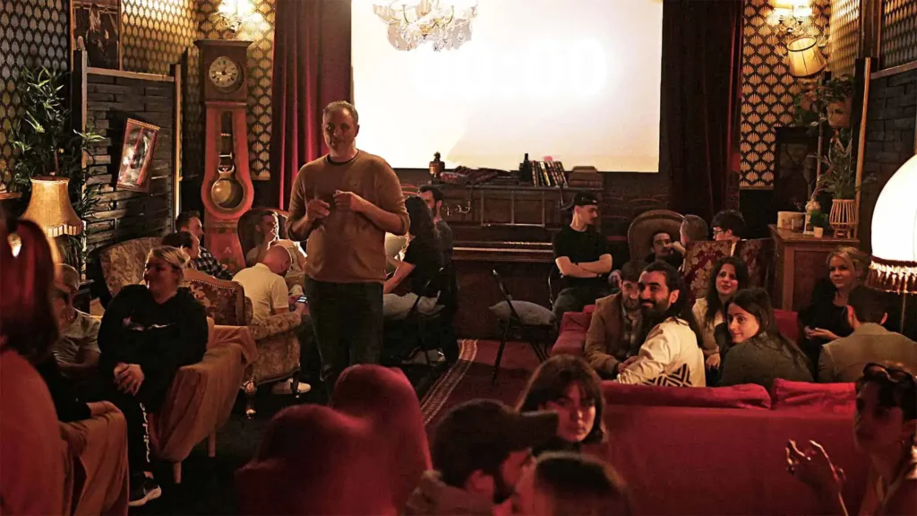 Un animateur guide une soirée murder party dans un salon vintage, avec un groupe de participants assis en demi-cercle, attentifs et impliqués dans l'activité.