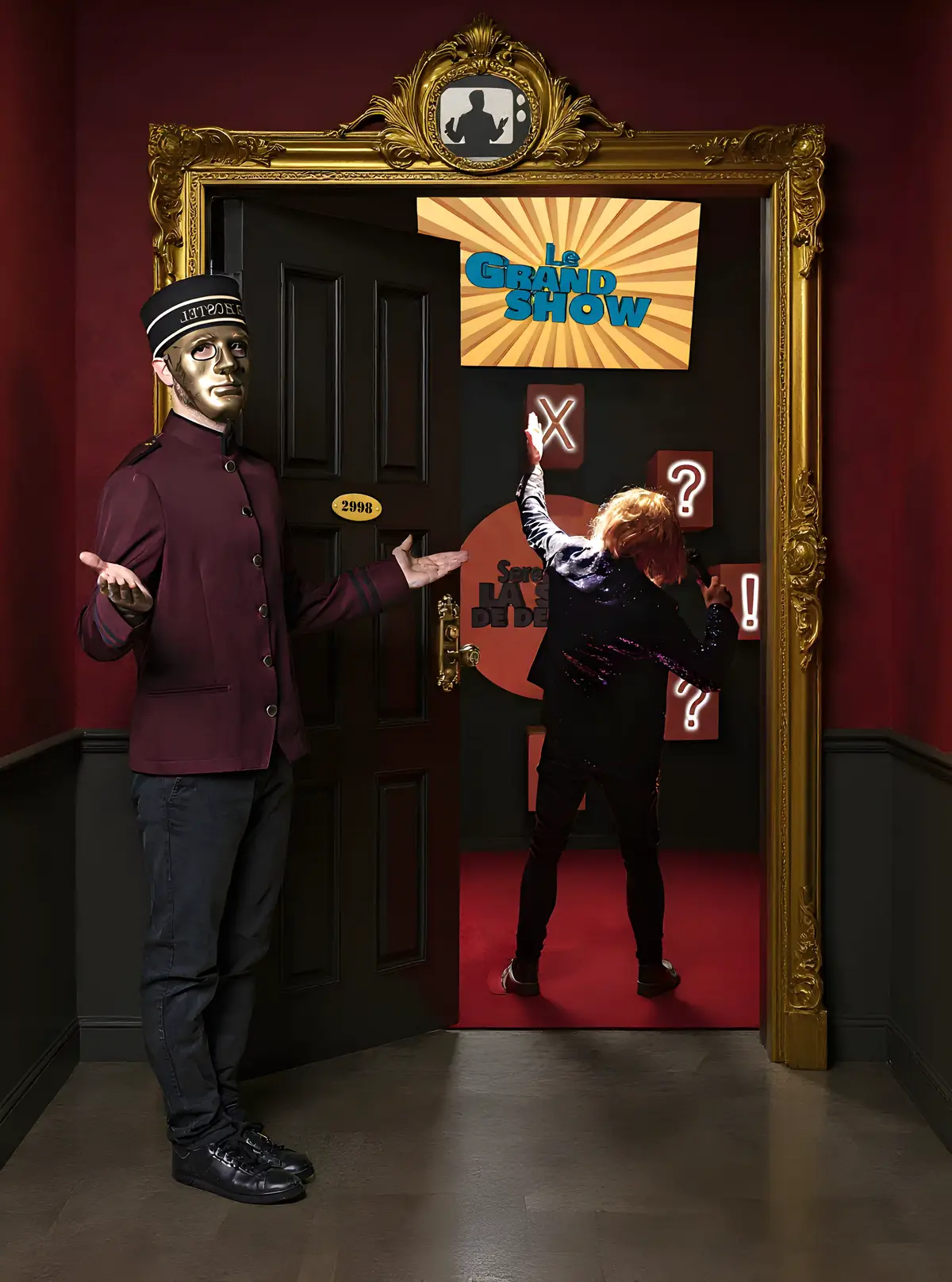 Un majordome à l'allure mystérieuse, vêtu d'un uniforme victorien et d'un masque doré, accueille un participant dans une pièce rouge décorée pour un escape game intitulé 'Le Grand Show."