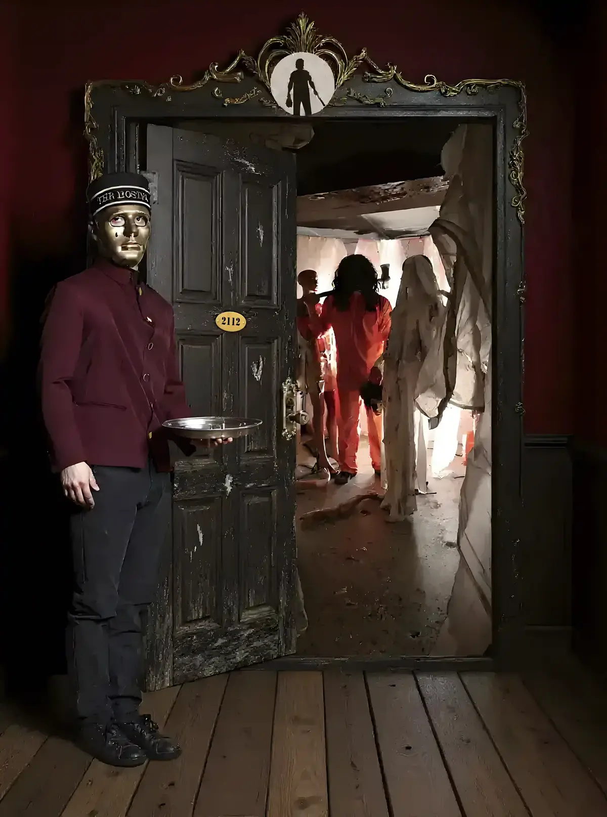 Un majordome au visage masqué, vêtu d'une veste pourpre et d'un chapeau noir, tient un plateau devant la porte de la chambre de l'hôtel The Hostel, révélant une scène d'horreur avec des personnages en combinaisons ensanglantées.