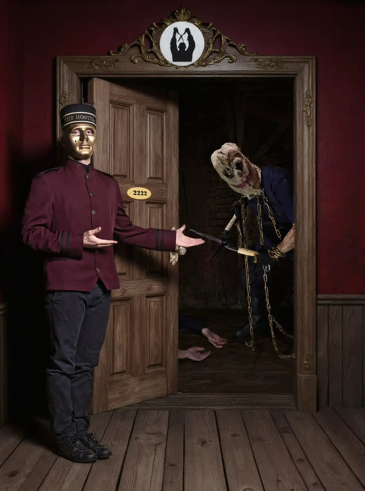 Un majordome au visage doré et en uniforme maroon ouvre une porte de chambre d'hôtel, révélant une scène horrifique : un homme enchaîné et défiguré a des cisailles dans la main.