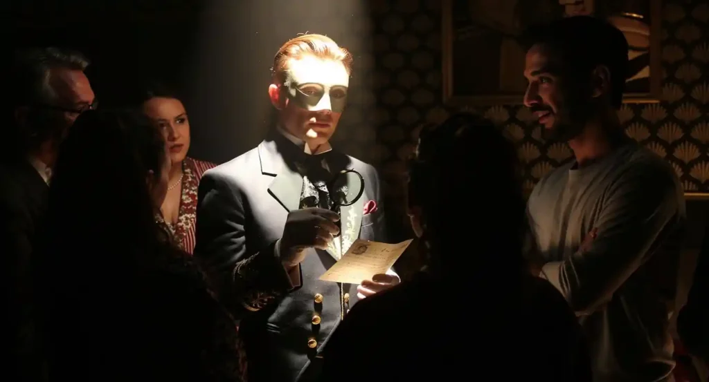 Un animateur masqué en costume victorien, tenant une loupe et une lettre ancienne, est mis en lumière par un éclairage théâtral. Autour de lui, un groupe de participants attentifs écoute dans une ambiance immersive et mystérieuse, évoquant une enquête ou une expérience interactive.