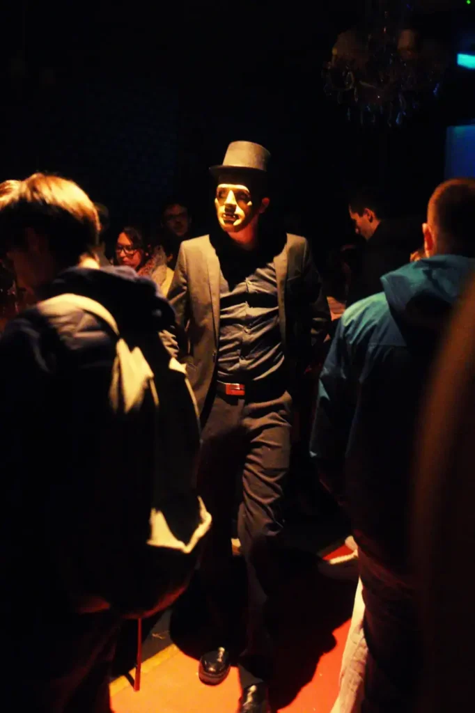 Personnage en costume élégant avec un chapeau haut-de-forme et un masque blanc, entouré de participants dans une ambiance tamisée - Scène immersive pour une Murder Party chez The Hostel