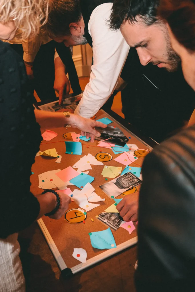 Groupe de personnes jouant à un escape game autour d'une table avec des indices, des photos et des accessoires colorés - Ambiance collaborative et immersive pour une activité chez The Hostel