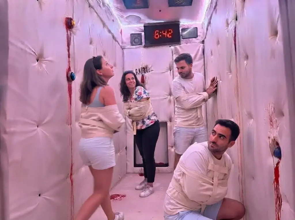 Groupe de quatre personnes dans une pièce thématique d'action game d'horreur, décorée avec des murs rembourrés et des traces de sang, en train de résoudre une énigme sous un chronomètre affichant 6:42