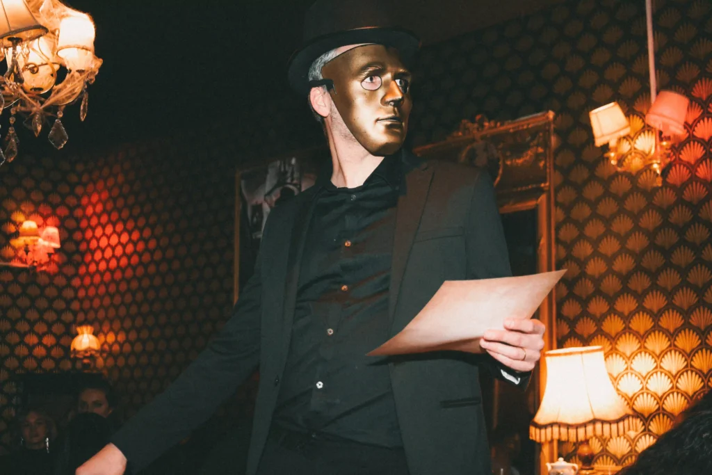 Un animateur en costume de majordome doré, portant un chapeau haut-de-forme et un masque doré, tient une feuille de papier dans une pièce décorée de motifs géométriques dorés et de luminaires vintage. Ambiance immersive pour un escape game ou une murder party.